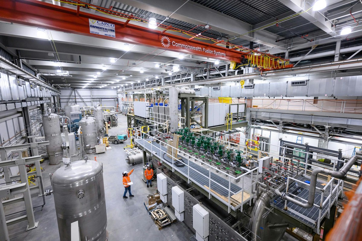 A large room with an orange overhead crane and workers in safety vests is filled with many silvery-gray ducts and green valves of cooling equipment.